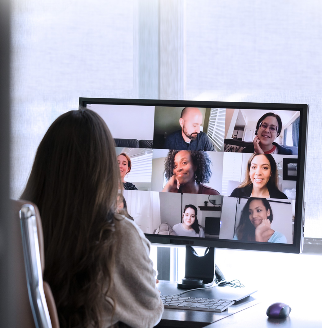 Žena sa dugom kosom sedi za stolom i učestvuje u video konferenciji na ekranu računara, gde su prikazana lica nekoliko kolega.