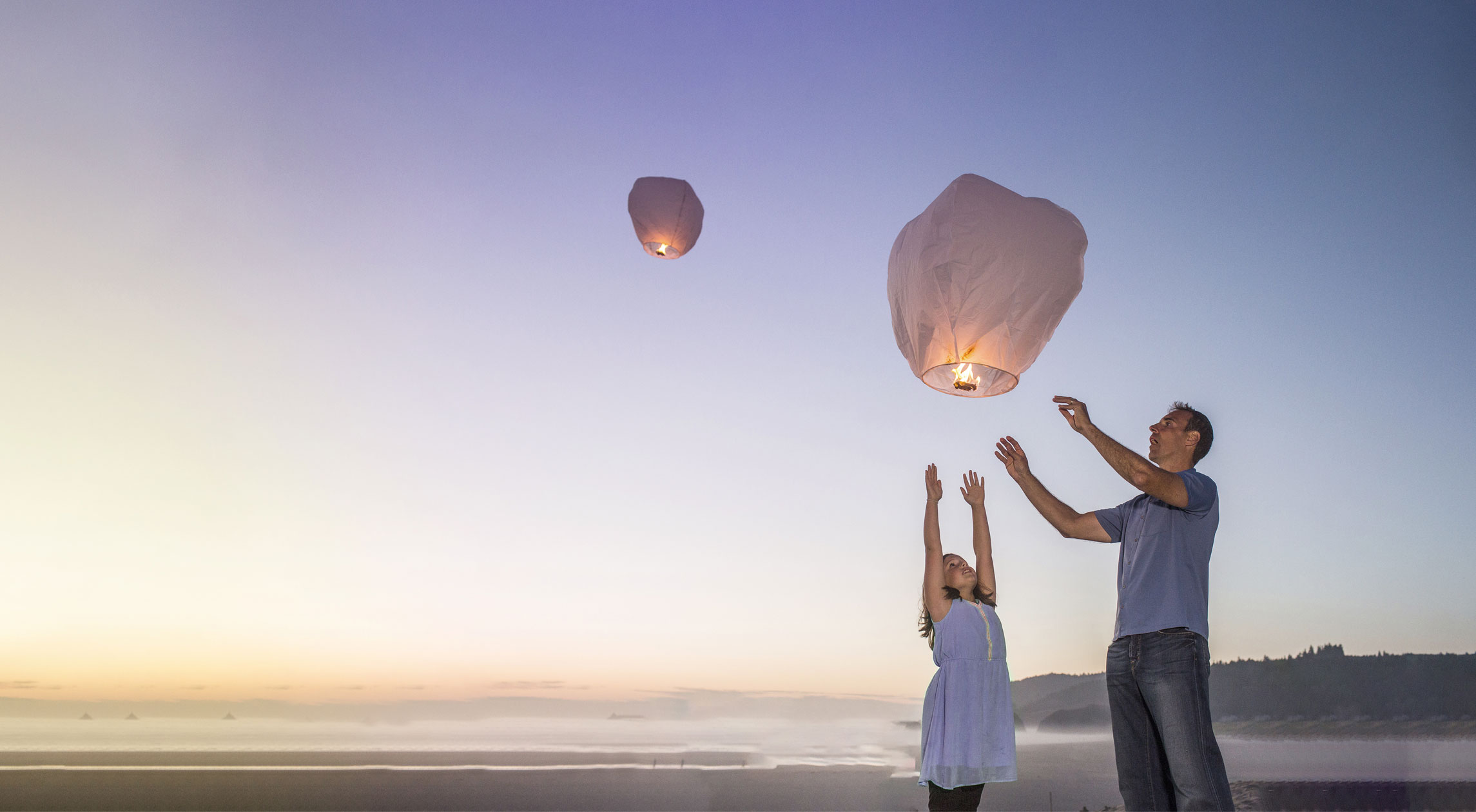Muškarac i devojčica na plaži puštaju svetleće lampione u nebo pri zalasku sunca, stvarajući magičnu atmosferu.