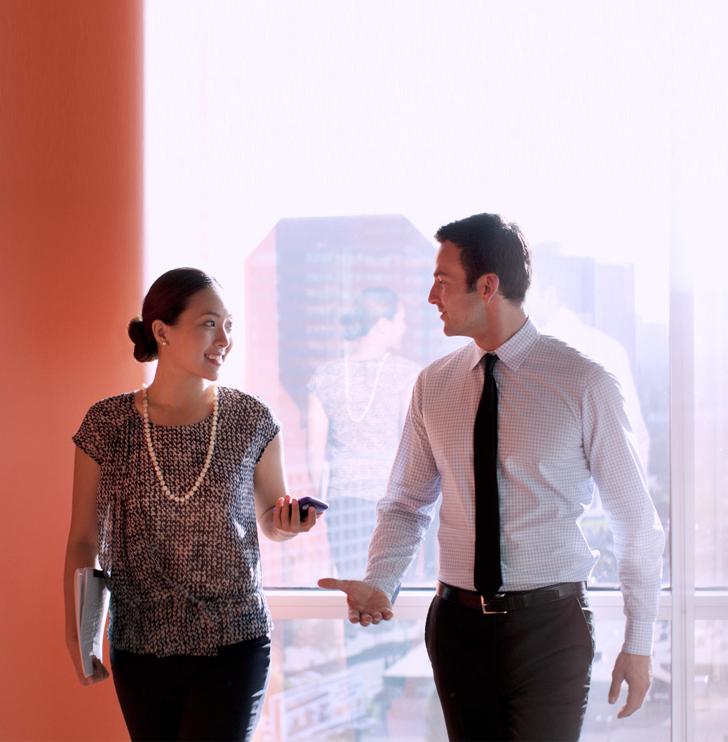 Poslovni čovek i žena hodaju kroz modernu kancelariju sa velikim prozorima, razgovarajući i deleći ideje u profesionalnom okruženju.