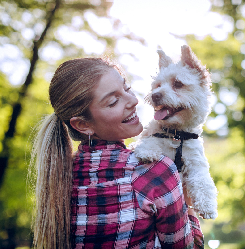 Mlada žena svezane kose u kariranoj košulji nasmejano drži svog psa dok stoje napolju u prirodi, okruženi zelenilom.