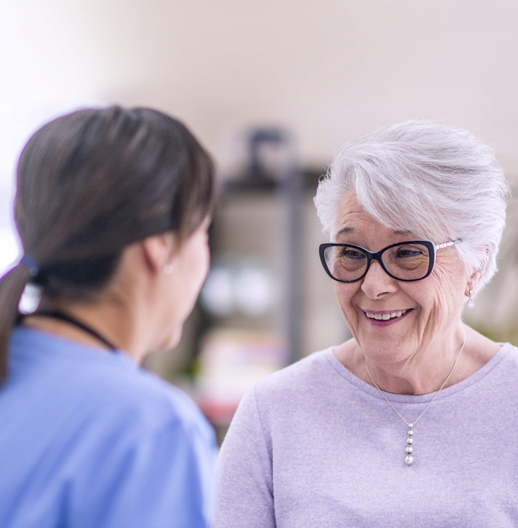 Mlada medicinska radnica razgovara sa nasmejanom starijom ženom s naočarima, pružajući podršku i brigu u zdravstvenom okruženju.