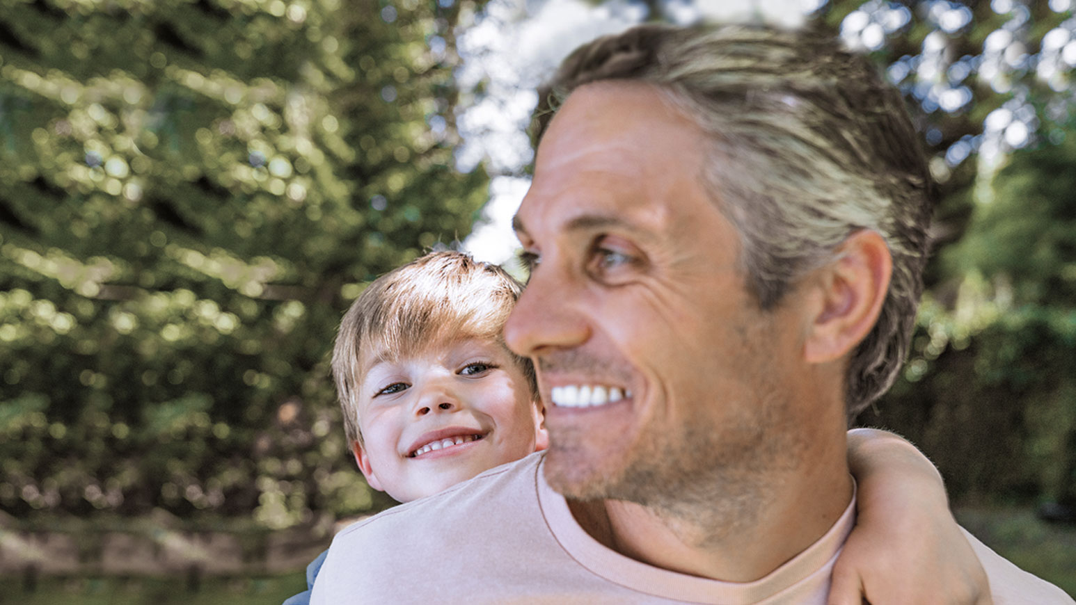 Nasmejani muškarac sa sedom kosom nosi dete na leđima, dok zajedno uživaju u prirodi