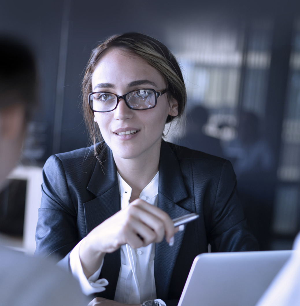 Poslovna žena sa naočarima i u tamnom sakou sedi za stolom sa laptopom, nasmejana i fokusirana.
