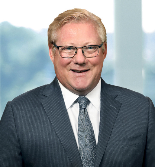 Portret nasmejanog muškarca u tamnoplavom odelu, beloj košulji i kravati, sa naočarima, pozira ispred modernog kancelarijskog prozora.