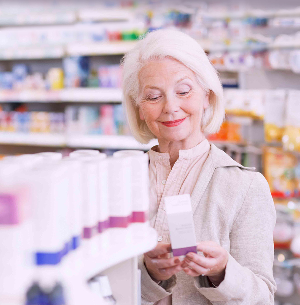 Starija žena sa sedom kosom i osmehom stoji u apoteci, držeći kutiju proizvoda dok pregleda police sa lekovima i suplementima
